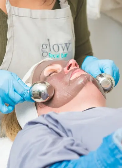 Ice globes rolled over a client's masked face by an esthetician in a glow facial bar apron