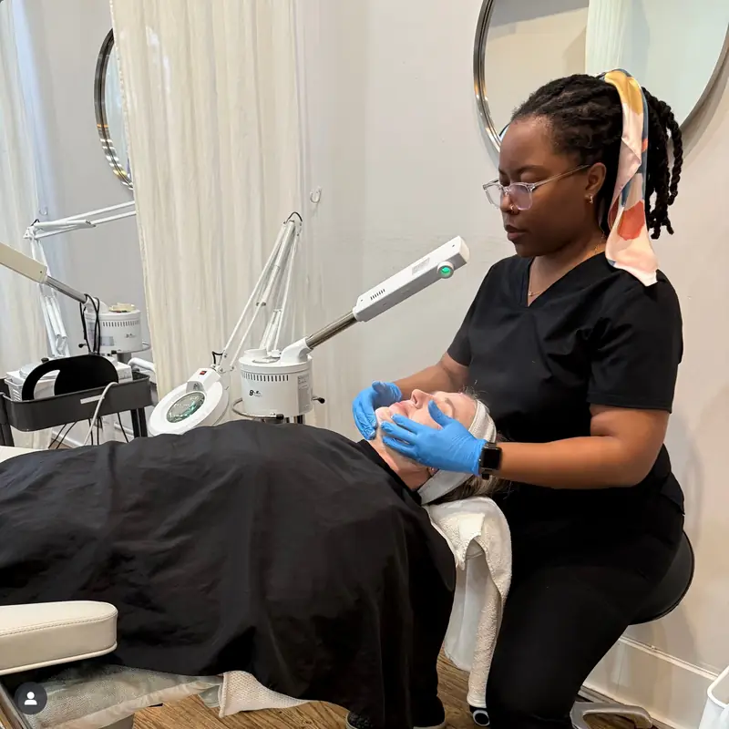 An esthetician in black scrubs working at a steamer station over a reclined client